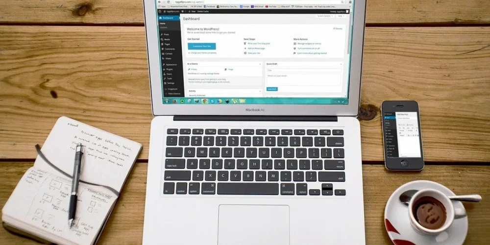 Srebrny laptop MacBook Air z otwartym panelem administracyjnym WordPress na ekranie, leżący na drewnianym stole obok notatnika z odręcznymi szkicami, smartfona i filiżanki kawy.